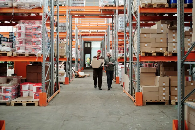 Two warehouse workers carry a cardboard box through an aisle of shelving and stacked cartons.